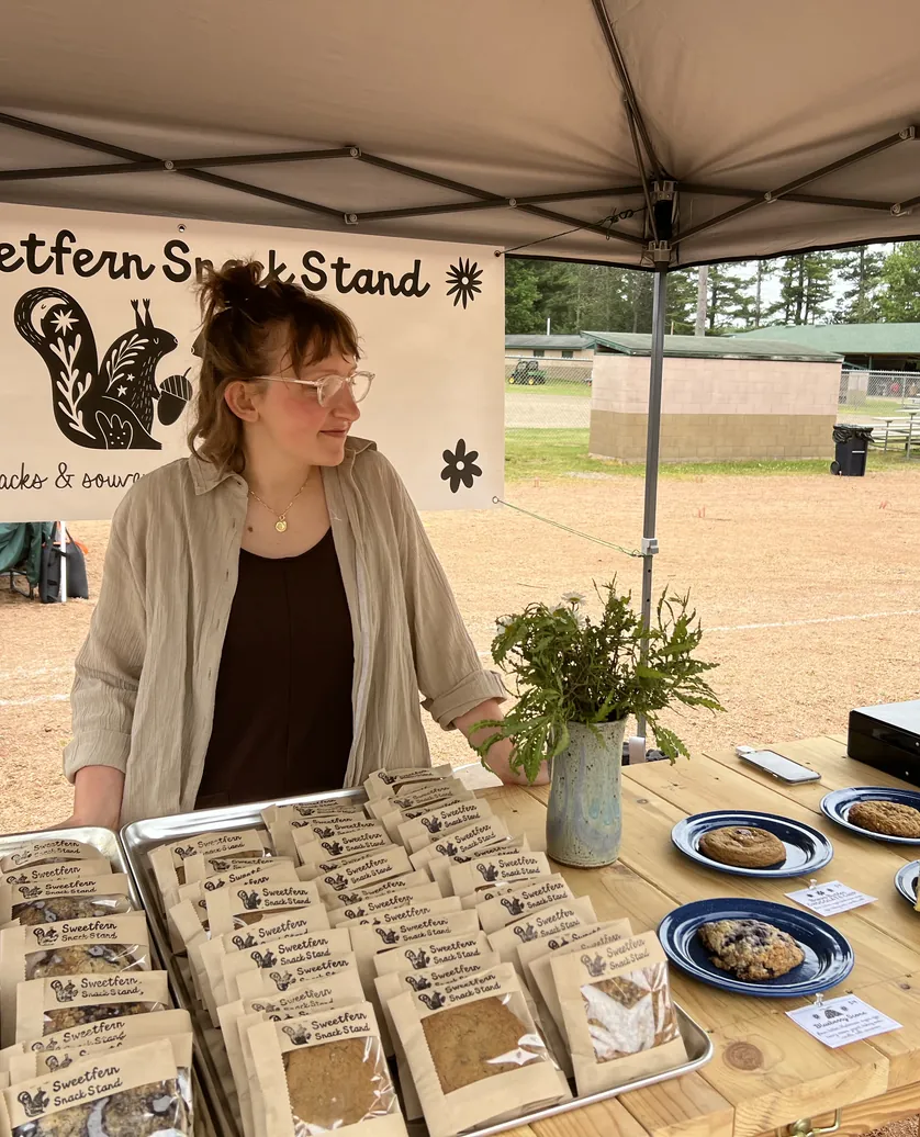 Kaitlin behind the Sweetfern Snack Stand table at a farmers market, bagged bakes laid out in front of the painted sign.