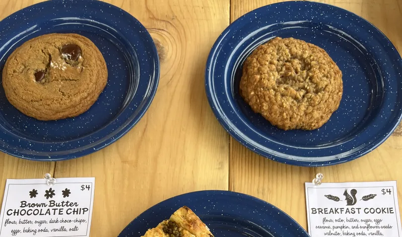 Two cookies on blue enamel plates at a market table, beside Kaitlin’s handwritten cards for the brown butter chocolate chip and the breakfast cookie.